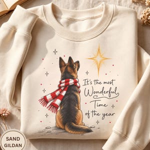 May include: A sand-colored crewneck sweatshirt featuring a graphic of a German Shepherd dog wearing a red and white plaid scarf, looking up at a glowing star. The text reads, It's the most Wonderful Time of the year, surrounded by small stars.