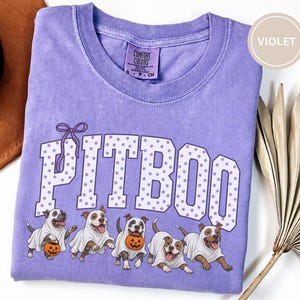 May include: A violet short-sleeved t-shirt with a crew neckline and a Comfort Colors label. The graphic features the text PITBOO in white with purple polka dots and a purple bow, accompanied by five illustrations of dogs wearing ghost costumes.