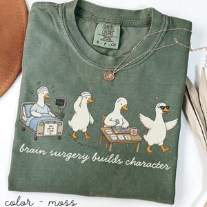 Op de afbeelding: Mosgroen Comfort Colors T-shirt met een cartoon van eenden in verschillende situaties. De tekst "brain surgery builds character" staat eronder. Een gouden ketting is ook zichtbaar. Maat S.