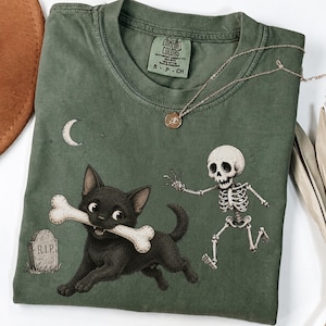 May include: An olive green crew neck t-shirt featuring a graphic of a black cat carrying a bone in its mouth, chased by a small skeleton. A tombstone marked R.I.P. and a crescent moon are visible. The shirt has a Comfort Colors brand label.