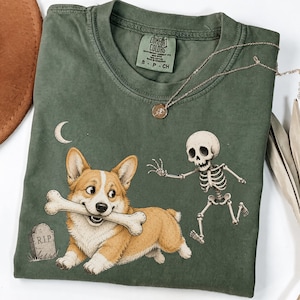 May include: An olive green t-shirt featuring a graphic of a corgi holding a bone in its mouth, a running skeleton, a crescent moon, and a tombstone labelled R.I.P. The shirt has a crew neckline and a Comfort Colors brand label on the inner collar.