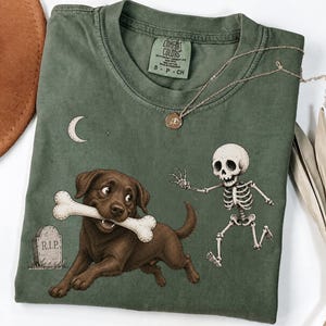 Pode incluir: Uma t-shirt verde-azeitona com um gráfico de um cão castanho a segurar um osso grande na boca, um esqueleto a correr, uma lápide com o texto R.I.P. e uma lua crescente. A etiqueta Comfort Colors indica o tamanho S, 100% algodão.