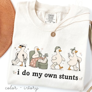 May include: Ivory-colored Comfort Colors t-shirt featuring a whimsical graphic of cartoon ducks with leg casts, each in a different pose. The text "i do my own stunts" is below the ducks. A gold necklace and a brown hat are also visible.