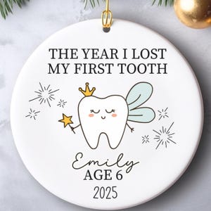 May include: White ceramic Christmas ornament with gold hanging loop. The ornament features the text "THE YEAR I LOST MY FIRST TOOTH" and a cartoon tooth fairy with a crown and wand. The name "Emily", age 6, and the year 2025 are also printed on the ornament.