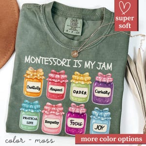 May include: Moss green t-shirt with the phrase "MONTESSORI IS MY JAM" and colorful jam jar graphics. Each jar has a different label, including "Creativity," "Respect," and "Joy." The shirt has a "super soft" label.