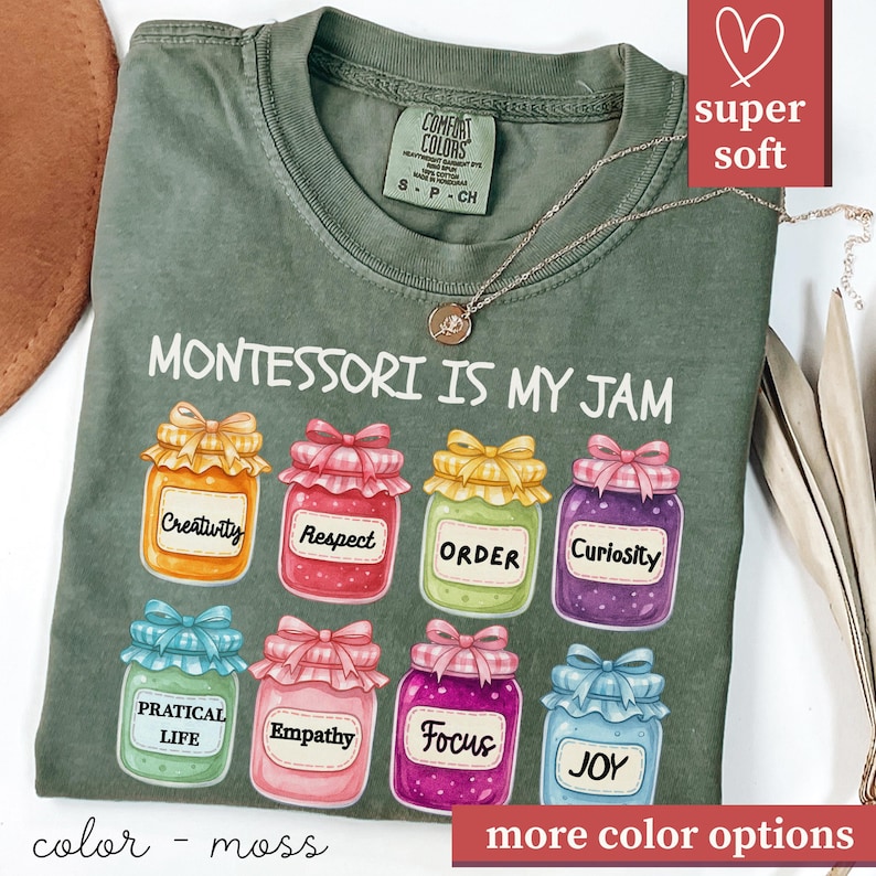 May include: Moss green t-shirt with the phrase "MONTESSORI IS MY JAM" and colorful jam jar graphics. Each jar has a different label, including "Creativity," "Respect," and "Joy." The shirt has a "super soft" label.