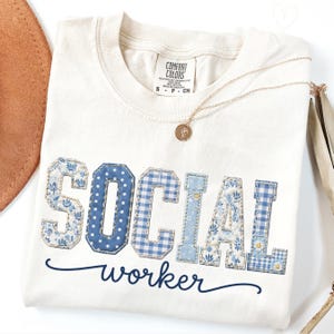 以下が含まれることがあります： クリーム色のComfort Colors Tシャツ。青色のパッチワーク文字で「SOCIAL WORKER」と書かれています。文字は花柄、水玉模様、ギンガムチェック柄です。金のネックレスと茶色の帽子が見えます。