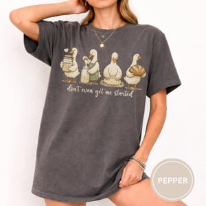 May include: Dark grey t-shirt with a cartoon of four ducks, each with baking items. The text "don't even get me started" is below the ducks. The shirt has a relaxed, oversized fit.