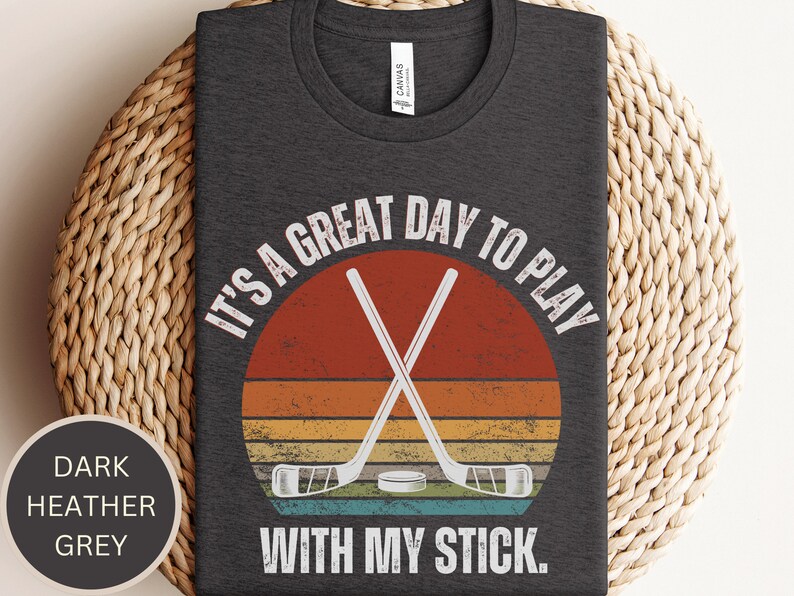 K&ouml;nnte beinhalten: Dunkelgraues T-Shirt mit einem Vintage-Grafik von gekreuzten Hockeyschl&auml;gern &uuml;ber einem Puck. Der Text lautet "Es ist ein gro&szlig;artiger Tag, um mit meinem Schl&auml;ger zu spielen."