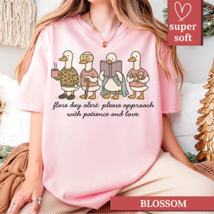 May include: A light pink t-shirt featuring a cartoon illustration of four ducks, each engaged in different activities. The text "flare day alert: please approach with patience and love" is printed below the ducks. The shirt also has the words "super soft" and "BLOSSOM".