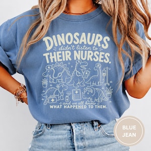 May include: A blue crewneck t-shirt featuring a cream-colored graphic of various dinosaurs dressed as medical professionals. The text reads: DINOSAURS didn't listen to THEIR NURSES. and we all know WHAT HAPPENED TO THEM. The design includes medical icons.