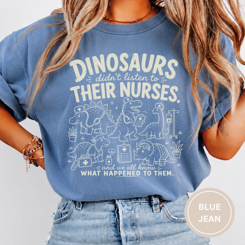 May include: A blue crewneck t-shirt featuring a cream-colored graphic of various dinosaurs dressed as medical professionals. The text reads: DINOSAURS didn't listen to THEIR NURSES. and we all know WHAT HAPPENED TO THEM. The design includes medical icons.