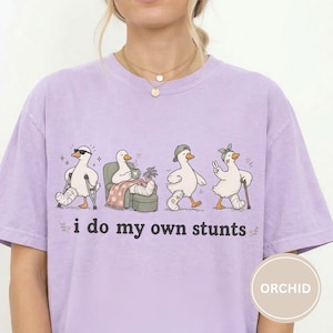May include: A light purple t-shirt featuring four cartoon geese with various injuries. The geese are depicted with casts, crutches, and bandages. The text "i do my own stunts" is printed below the geese in a playful font.