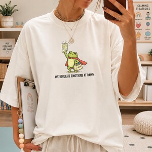 May include: White t-shirt featuring a cartoon frog superhero holding a chart and a shopping bag. The frog wears a red cape. Text on the shirt reads "WE REGULATE EMOTIONS AT DAWN". Additional text on the background includes "ALL FEELINGS ARE OKAY".