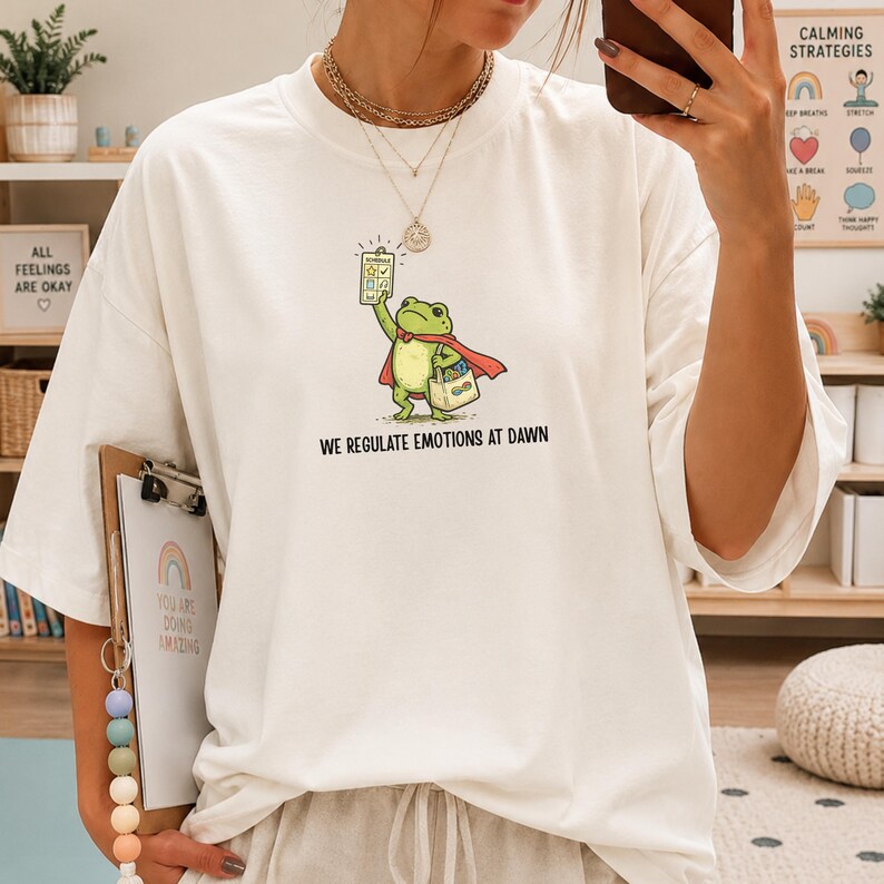 May include: White t-shirt featuring a cartoon frog superhero holding a chart and a shopping bag. The frog wears a red cape. Text on the shirt reads "WE REGULATE EMOTIONS AT DAWN". Additional text on the background includes "ALL FEELINGS ARE OKAY".