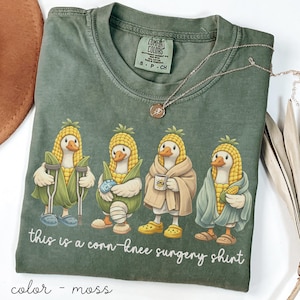 Könnte beinhalten: Moosgrünes T-Shirt mit einer Grafik von vier Enten, die als Maiskolben verkleidet sind, jede mit einer anderen Verletzung oder einem anderen Erholungsgegenstand. Der Text "this is a corn-knee surgery shirt" steht darunter.