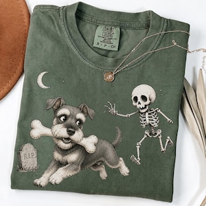 Puede incluir: Una camiseta de manga corta color verde oliva con un gráfico de un perro schnauzer sosteniendo un hueso en la boca, un esqueleto corriendo, una luna creciente y una lápida con el texto R.I.P. La camiseta tiene una etiqueta Comfort Colors en el cuello.