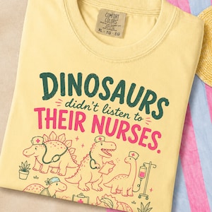 May include: A pale yellow t-shirt featuring the dark green and pink text "DINOSAURS didn't listen to THEIR NURSES." The graphic includes illustrations of four dinosaurs wearing nurse caps, with one holding a clipboard and another near an IV pole.