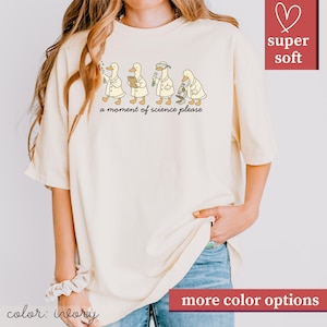 May include: Ivory-colored t-shirt featuring a whimsical illustration of four ducks in lab coats, each engaged in a different scientific activity. The phrase "a moment of science please" is printed below the ducks. The shirt has a relaxed fit and is labeled "super soft".
