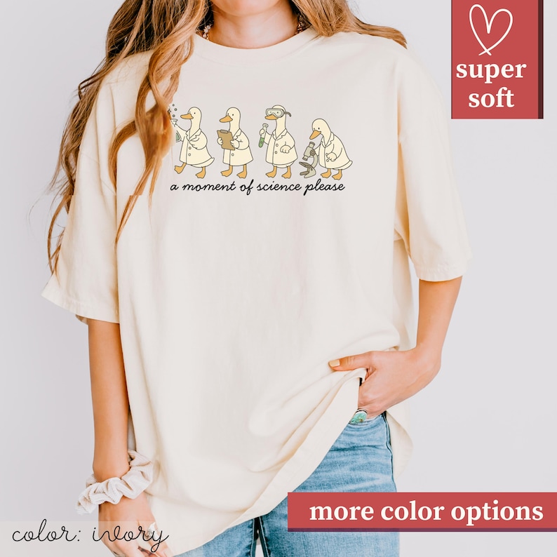 May include: Ivory-colored t-shirt featuring a whimsical illustration of four ducks in lab coats, each engaged in a different scientific activity. The phrase "a moment of science please" is printed below the ducks. The shirt has a relaxed fit and is labeled "super soft".