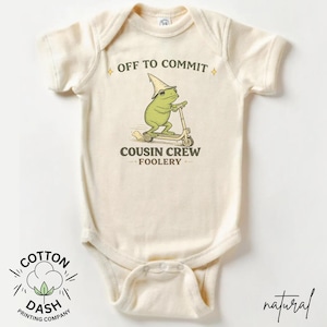 K&ouml;nnte beinhalten: Cremefarbener Baby-Body mit einer skurrilen Frosch-Grafik, die einen Roller f&auml;hrt und einen spitzen Hut tr&auml;gt. Der Text lautet "OFF TO COMMIT" und "COUSIN CREW FOOLERY". Das Logo der Cotton Dash Printing Company befindet sich unten.