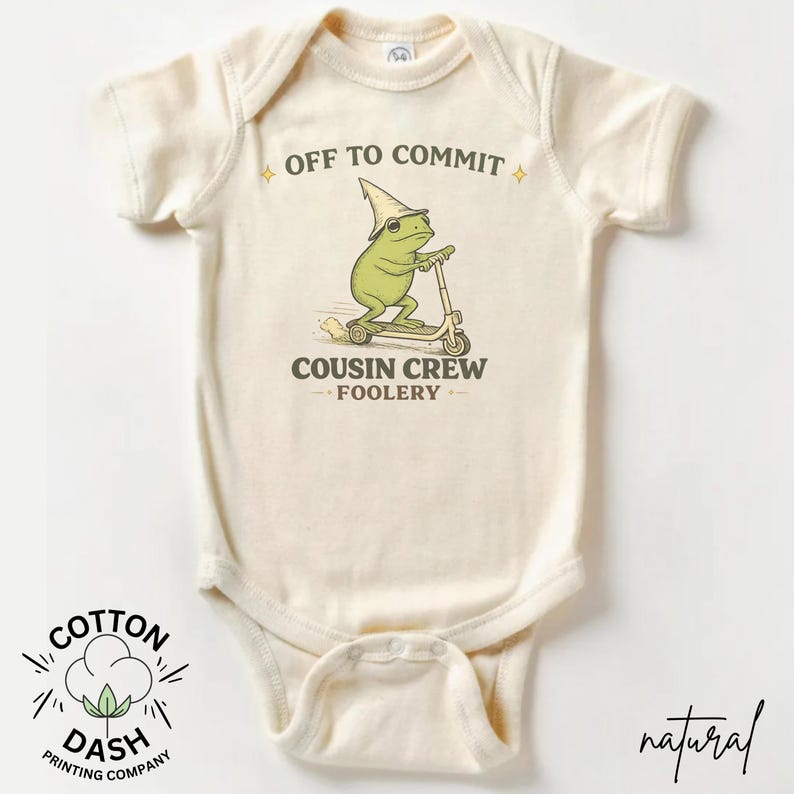 K&ouml;nnte beinhalten: Cremefarbener Baby-Body mit einer skurrilen Frosch-Grafik, die einen Roller f&auml;hrt und einen spitzen Hut tr&auml;gt. Der Text lautet "OFF TO COMMIT" und "COUSIN CREW FOOLERY". Das Logo der Cotton Dash Printing Company befindet sich unten.