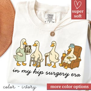 以下が含まれることがあります： アイボリー色のTシャツ。様々な回復段階の鴨のユーモラスなグラフィックが特徴で、氷嚢をしている鴨もいます。「in my hip surgery era」の文字が下にあります。Tシャツには「super soft」の文字もあります。