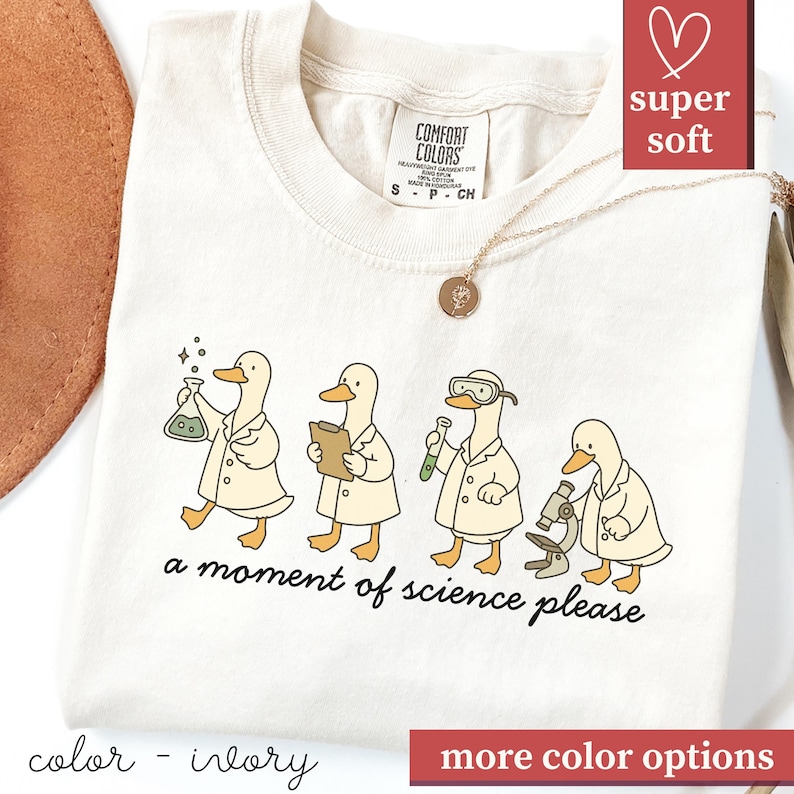 May include: Ivory-colored t-shirt featuring a whimsical illustration of four ducks in lab coats, each with scientific tools. The text "a moment of science please" is below the ducks. The shirt has a crew neck and short sleeves.