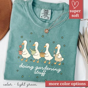 May include: Light green t-shirt featuring a graphic of four ducks engaged in gardening activities, each with a different tool. The phrase "doing gardening stuff" is printed below. The shirt has a crew neck and short sleeves. The words "super soft" are in a red box.