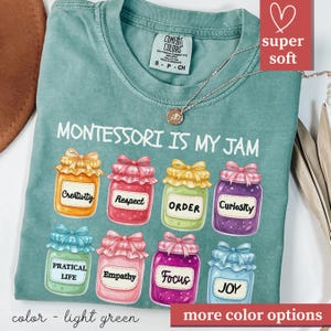 May include: Light green t-shirt with the text "MONTESSORI IS MY JAM" and illustrations of jam jars labeled with words like "Creativity" and "Joy." The shirt has a crew neck and short sleeves. A silver necklace with a pendant is also visible.