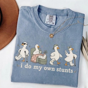 May include: A blue Comfort Colors t-shirt featuring a graphic of four ducks with leg casts, each in a different pose. The text "i do my own stunts" is below the ducks. A gold necklace is draped across the shirt.