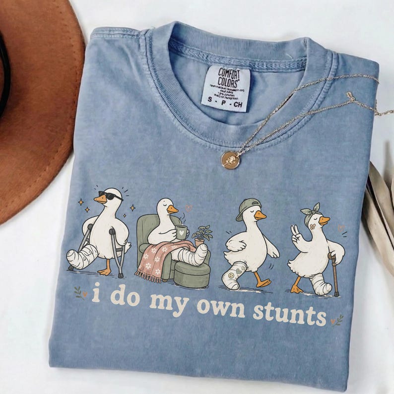 May include: A blue Comfort Colors t-shirt featuring a graphic of four ducks with leg casts, each in a different pose. The text "i do my own stunts" is below the ducks. A gold necklace is draped across the shirt.