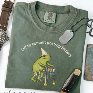 May include: Sage green Comfort Colors t-shirt with the phrase "Off to commit post-op foolery." A cartoon frog wearing a hat and knee brace uses a walker. A dog tag and a pocket knife are also visible.