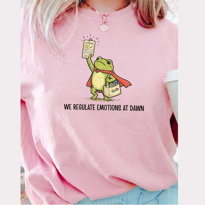 May include: Pink sweatshirt featuring a cartoon frog superhero holding a schedule and a shopping bag. The frog wears a red cape and the text "WE REGULATE EMOTIONS AT DAWN" is printed below the frog.