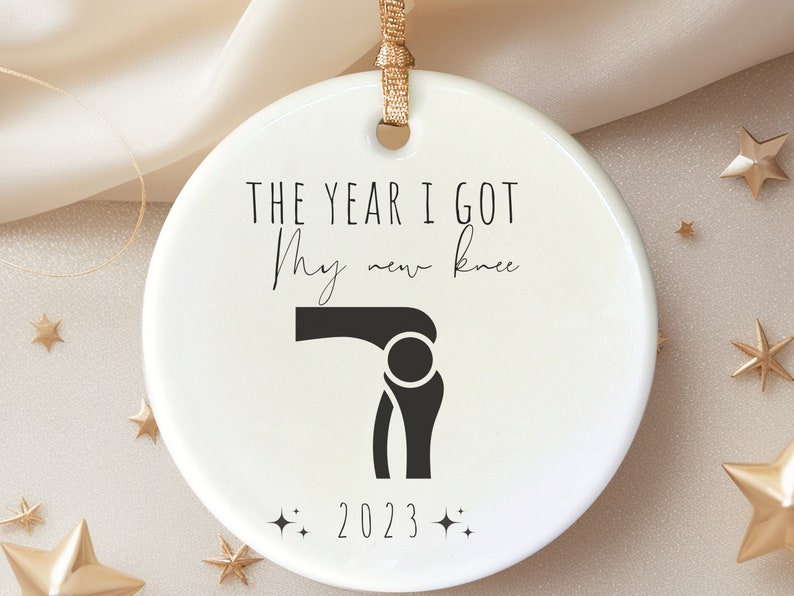 Op de afbeelding: Witte kerstbal van keramiek met zwarte tekst die "Het jaar dat ik mijn nieuwe knie kreeg" luidt en een zwart silhouet van een knie. Het jaar 2023 staat ook op de kerstbal gedrukt.