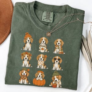 Könnte beinhalten: Ein olivgrünes T-Shirt mit Rundhalsausschnitt und einem Grafikdruck von neun Beagle-Welpen in verschiedenen herbstlichen Outfits und Accessoires, darunter Kürbisse, Schals, Mützen und Blumenkränze. Das Shirt hat ein Comfort Colors Etikett.