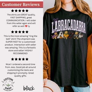 May include: A black t-shirt featuring the text LABRACADABRA in white polka-dot letters above a graphic of six Labrador dogs dressed in various Halloween costumes, including a witch, a ghost, a pumpkin, a vampire, a skeleton, and a mummy.