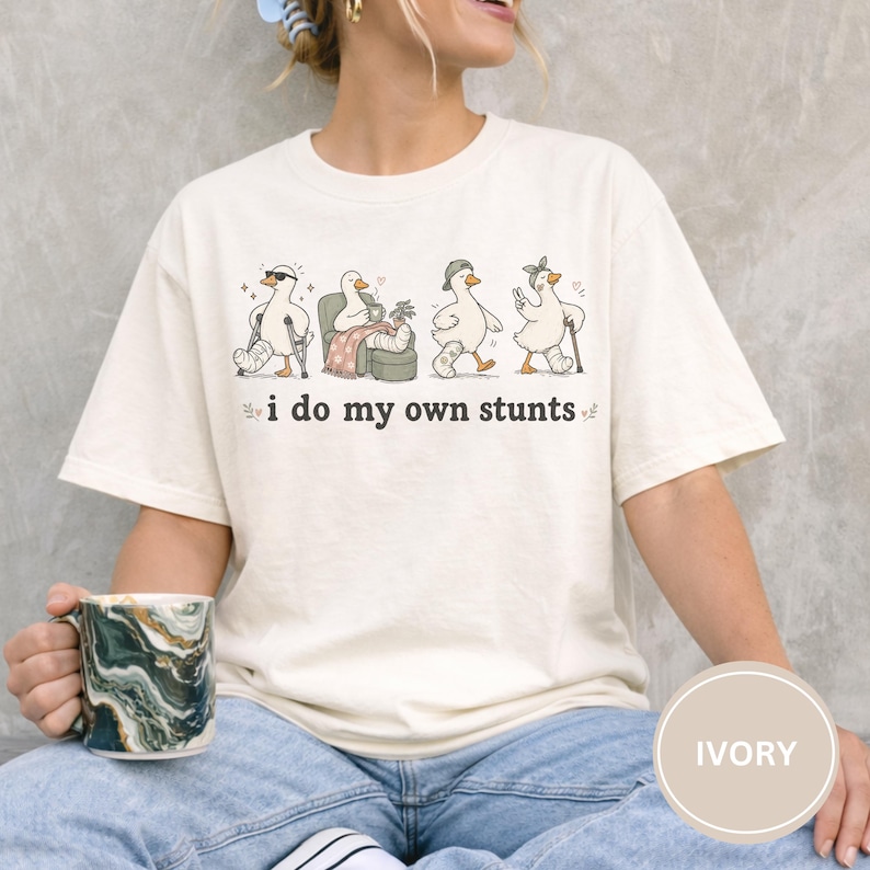 May include: Ivory-colored t-shirt featuring a whimsical illustration of four geese in various states of injury, each with a different accessory. The text "i do my own stunts" is printed below the geese. The shirt is a casual, comfortable style.
