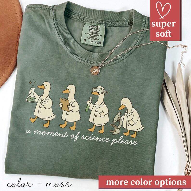 May include: Moss green Comfort Colors t-shirt featuring a line of cartoon ducks in lab coats, each with a different science tool. The text "a moment of science please" is below the ducks. A gold necklace is draped across the shirt.