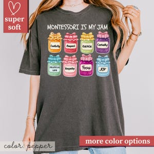 May include: A dark gray t-shirt with the text "MONTESSORI IS MY JAM" and colorful jar illustrations. Each jar has a different word: Creativity, Respect, Order, Curiosity, Practical Life, Empathy, Focus, and Joy. The shirt also has the words "super soft" and "more color options".
