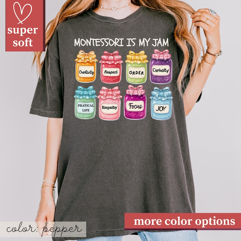 May include: A dark gray t-shirt with the text "MONTESSORI IS MY JAM" and colorful jar illustrations. Each jar has a different word: Creativity, Respect, Order, Curiosity, Practical Life, Empathy, Focus, and Joy. The shirt also has the words "super soft" and "more color options".