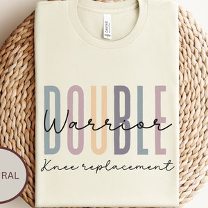 May include: A light beige t-shirt with the text "DOUBLE Warrior Knee Replacement" in colorful letters. The text is in a modern font style.