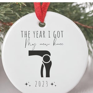 Op de afbeelding: Witte kerstbal van keramiek met zwarte tekst die "Het jaar dat ik mijn nieuwe knie kreeg" luidt en een zwart silhouet van een knie. Het jaar 2023 is onder de knie gedrukt.