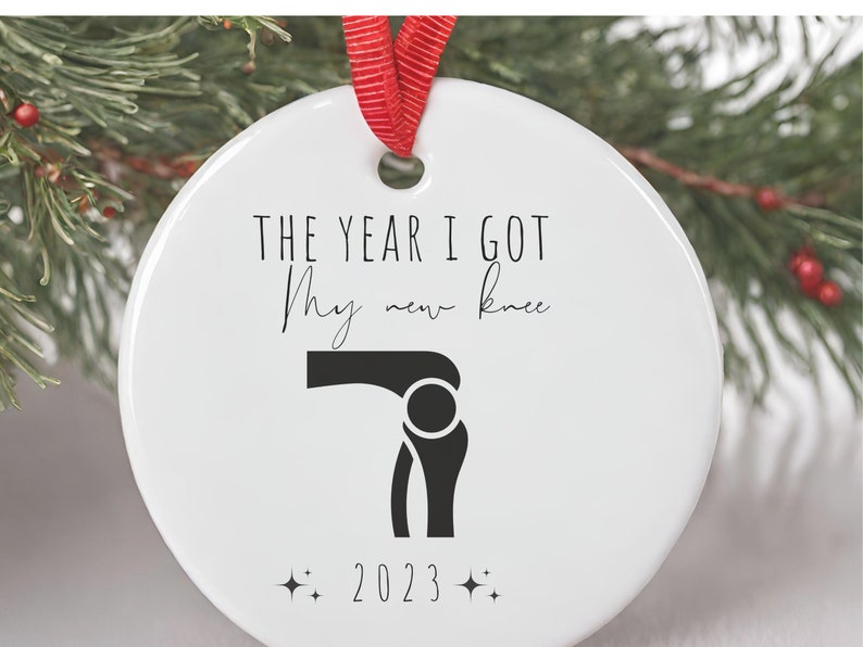 Op de afbeelding: Witte kerstbal van keramiek met zwarte tekst die "Het jaar dat ik mijn nieuwe knie kreeg" luidt en een zwart silhouet van een knie. Het jaar 2023 is onder de knie gedrukt.