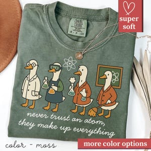 May include: A moss green t-shirt with a cartoon design of four ducks in lab coats, each holding a flask. The text "never trust an atom, they make up everything" is printed below the ducks. A gold necklace is draped across the shirt.