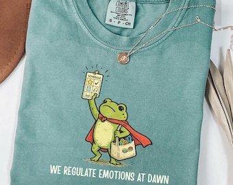 May include: Light green t-shirt with a cartoon frog superhero holding a schedule and a shopping bag. The text "WE REGULATE EMOTIONS AT DAWN" is printed below the frog. A gold necklace with a pendant is draped across the shirt.