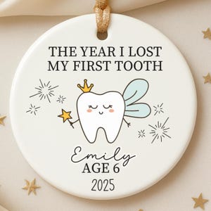 May include: A white ceramic ornament with a gold ribbon for hanging. The ornament features the text "THE YEAR I LOST MY FIRST TOOTH" and a cartoon tooth fairy with a crown and wand. The name "Emily", age 6, and the year 2025 are also included.