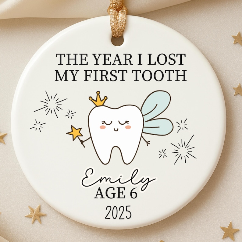 May include: A white ceramic ornament with a gold ribbon for hanging. The ornament features the text "THE YEAR I LOST MY FIRST TOOTH" and a cartoon tooth fairy with a crown and wand. The name "Emily", age 6, and the year 2025 are also included.