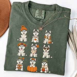Könnte beinhalten: Ein olivgrünes T-Shirt mit Rundhalsausschnitt und einem Grafikdruck von neun Husky-Welpen mit verschiedenen herbstlichen Accessoires wie Mützen, Schals und Kürbissen. Das Innenetikett am Hals zeigt den Text Comfort Colors S P CH.