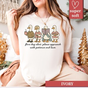 May include: Ivory-colored t-shirt featuring a whimsical illustration of four ducks, each engaged in different activities. The ducks are depicted with various accessories and clothing. The text "flare day alert: please approach with patience and love" is printed below the ducks.